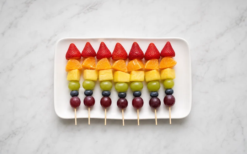 Rainbow fruit kabobs on wooden skewers with strawberries oranges pineapple grapes and blueberries on a white platter