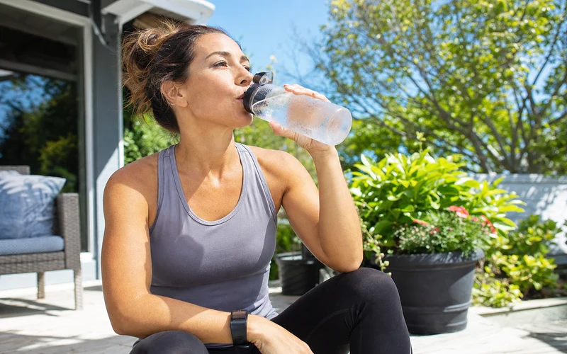Woman drinking cold water from a reusable bottle on a sunny summer patio