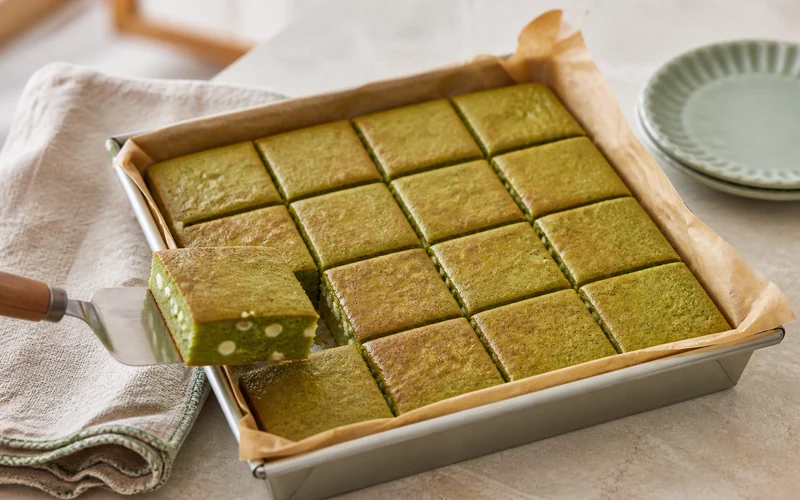 Sixteen chewy matcha mochi bars with white chocolate in a parchment-lined square pan.