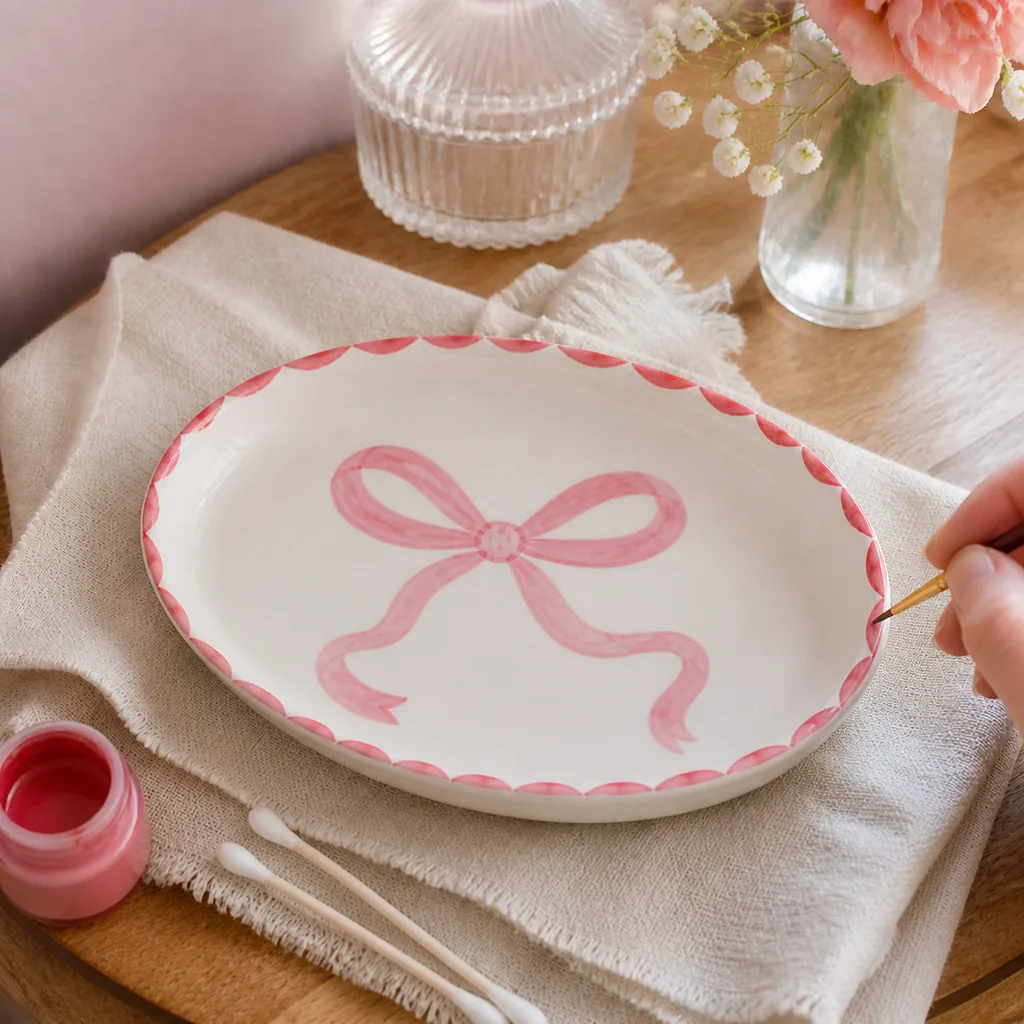 Red scalloped rim being painted around a white ceramic trinket dish with a dusty pink bow in the center.