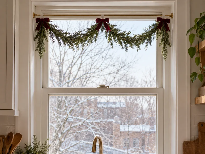 Light faux Christmas garland tied to a brass tension rod across a recessed kitchen window.
